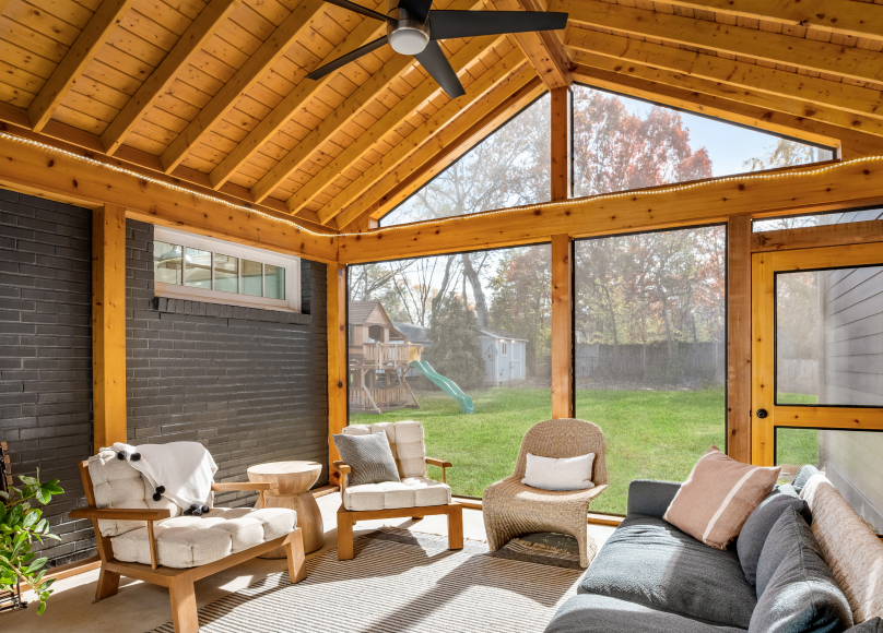 A beautifully designed screened-in porch with wooden beams, cozy seating, and a scenic backyard view.