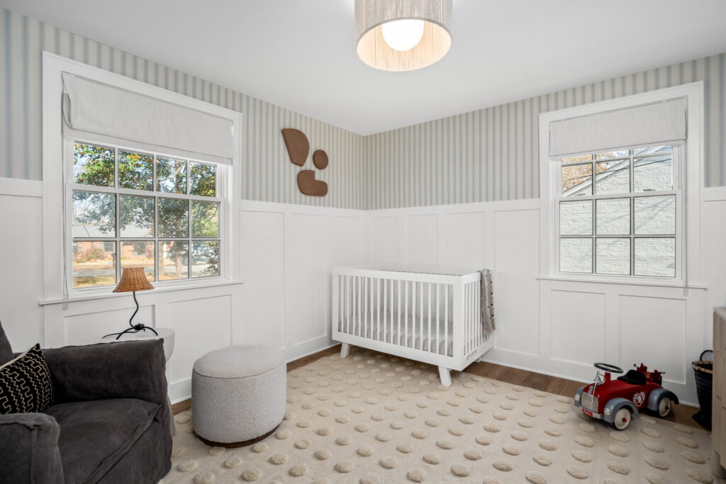 Children's room with soft neutral tones, a white crib, playful accents, and natural light streaming through double windows.